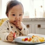 Adorable baby in a polka dot outfit enjoying fresh fruits in a high chair at home.