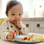 Adorable baby in a polka dot outfit enjoying fresh fruits in a high chair at home.