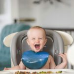 Happy baby laughing in a high chair during mealtime, wearing a blue bib.
