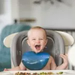 Happy baby laughing in a high chair during mealtime, wearing a blue bib.
